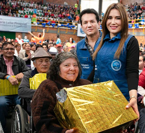 Dr. César Moreno and Dr. Ángela Oviedo handing a golden package to a person with a disability at an event, surrounded by a crowd in a coliseum.